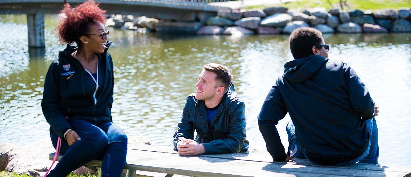 Three students sitting on a picnic table in front of Bear Lake at Oakland University.